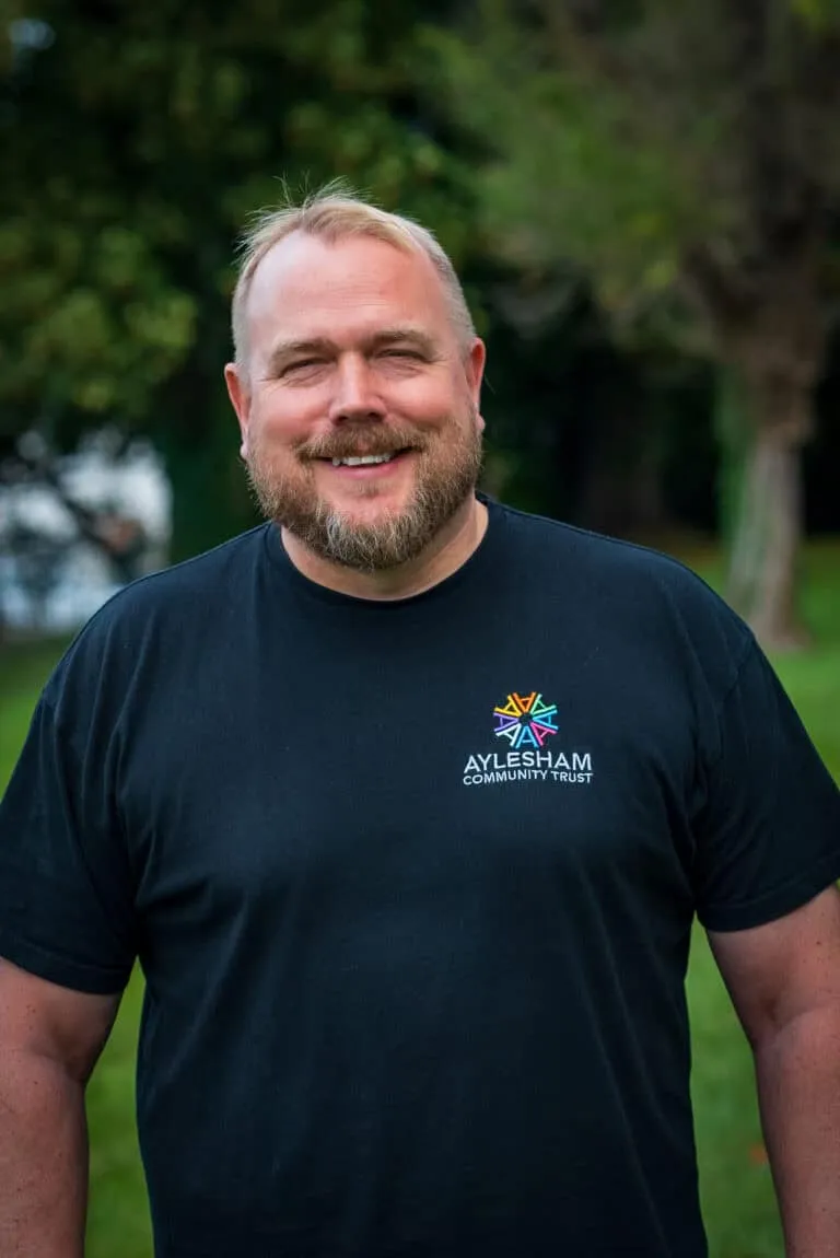 Community leader smiling outdoors, wearing a black Aylesham Community Trust t-shirt, promoting local initiatives and community development in Aylesham.