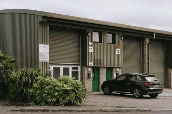 Modern industrial warehouse building with green doors and numbered units, surrounded by greenery, representing Aylesham Community Trust's facilities and local business hub.
