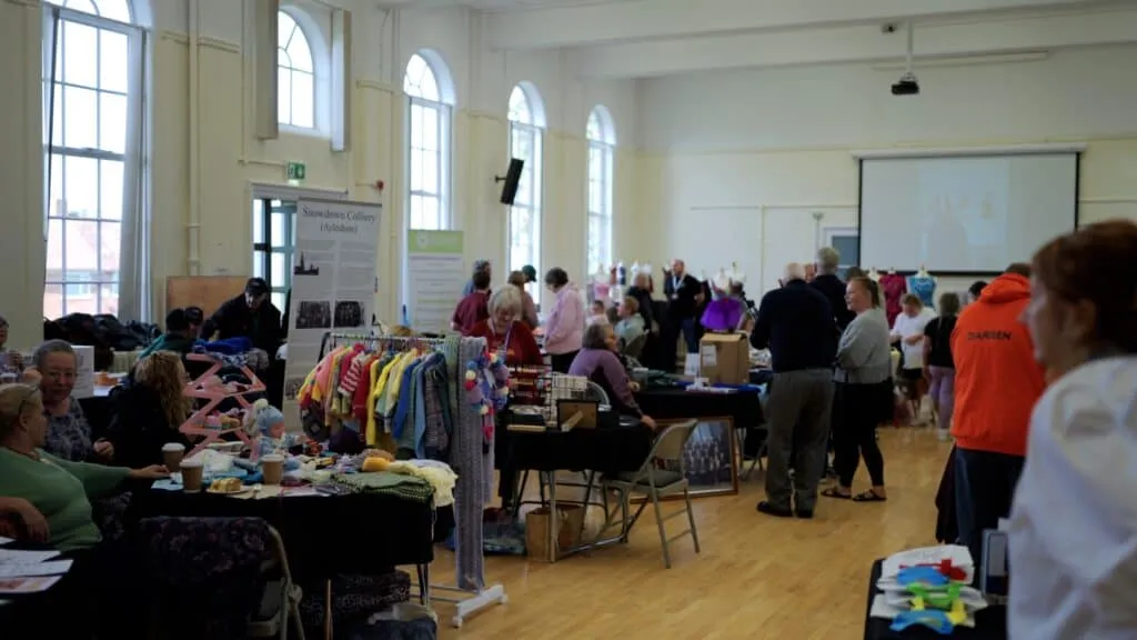 Community event at Aylesham Community Trust with local vendors showcasing crafts, knitting, and community activities in a welcoming hall setting.