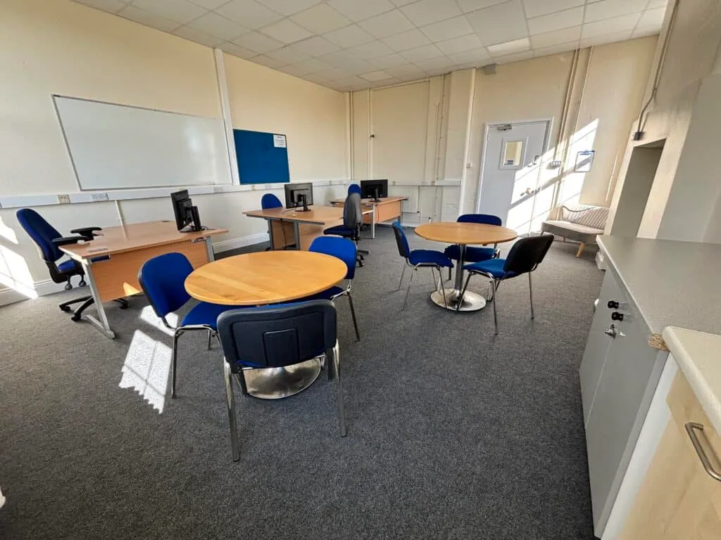 Bright community room with tables and chairs, computer stations, and natural sunlight, ideal for local events, meetings, and activities at Aylesham Community Trust.